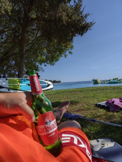 liegend am Strand mit einer Flasche Bier in der Hand im Hintergrund ein SUP und das Meer