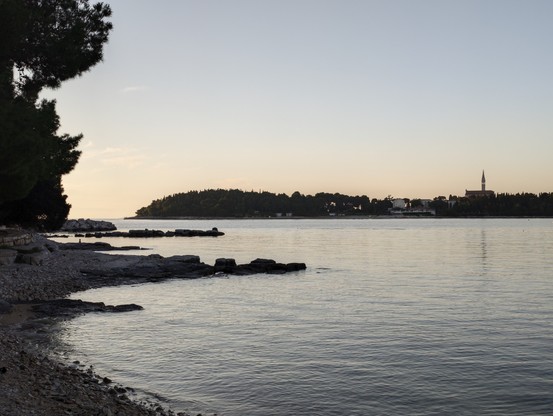 Strand mit kleinen und großen Steinen die ins Meer ragen, im Hintergrund eine Insel mit einer Kirche das ganze in Sonnenuntergangslicht getaucht